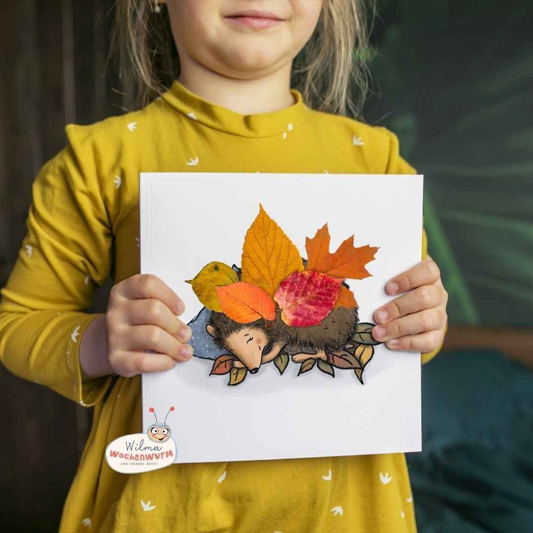 Kind in gelbem Pullover hält fertiges Igel Bastelprojekt mit bunten Herbstblättern hoch - Igel Nest basteln Wilma Wochenwurm